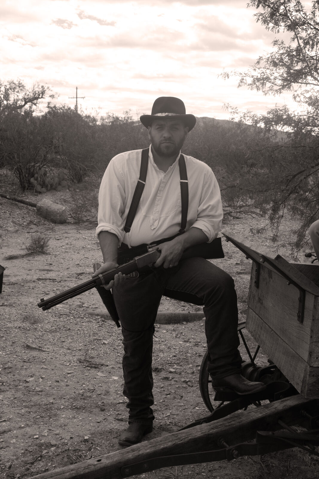 Man in a hat and suspenders holding a rifle beside a wagon