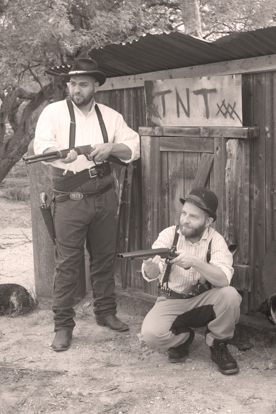 Two men with rifles in front of a weathered shed marked TNT
